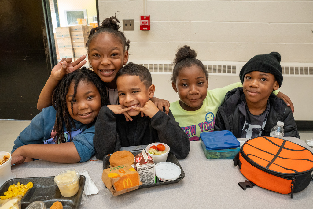Five students posing together at lunch table with their meal trays, smiling at camera with basketball lunchbox visible