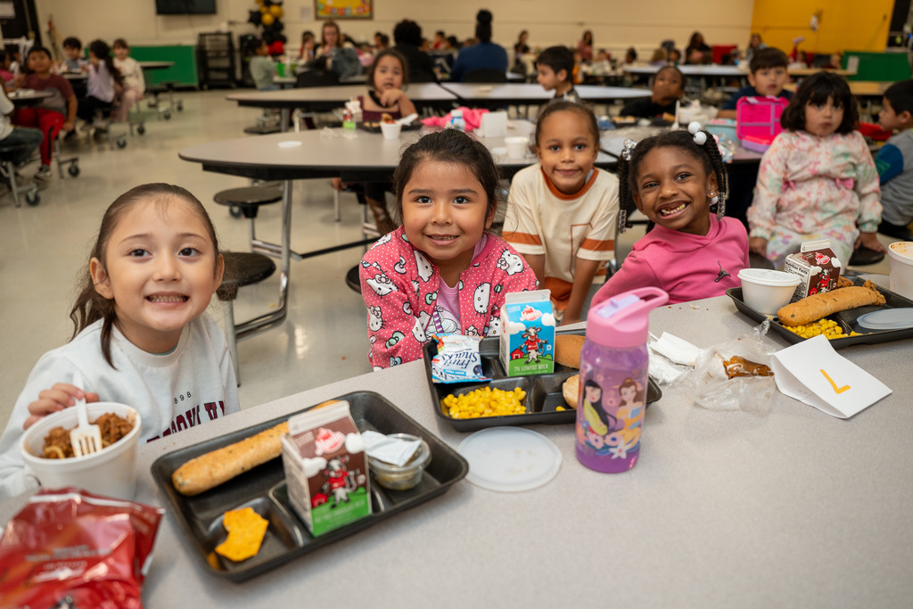 Four elementary students sitting together at lunch table smiling at camera with their lunch trays containing various menu items