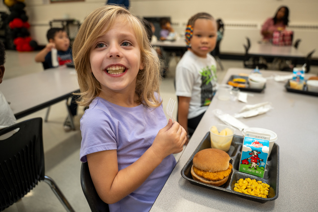 Young student in purple shirt giving thumbs up while sitting at lunch table with chicken sandwich, corn, and milk carton