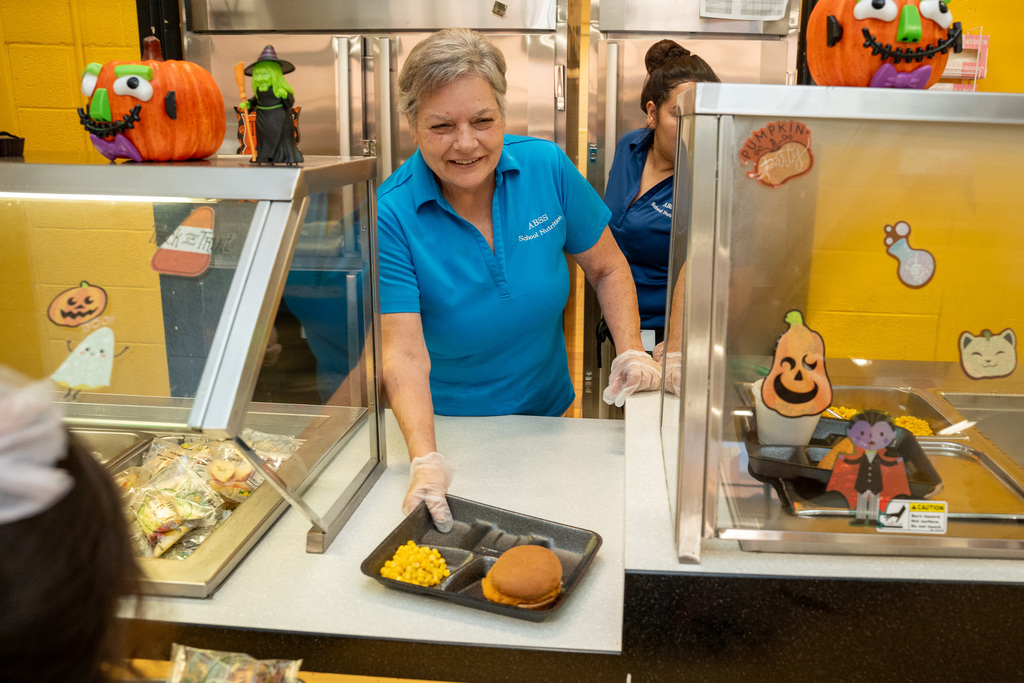 Cafeteria worker in blue ABSS uniform serving lunch through service window decorated with Halloween cutouts, placing chicken sandwich and corn on tray