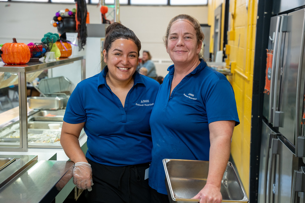 Two cafeteria staff members in blue ABSS School Nutrition uniforms standing together in kitchen with Halloween decorations visible in background