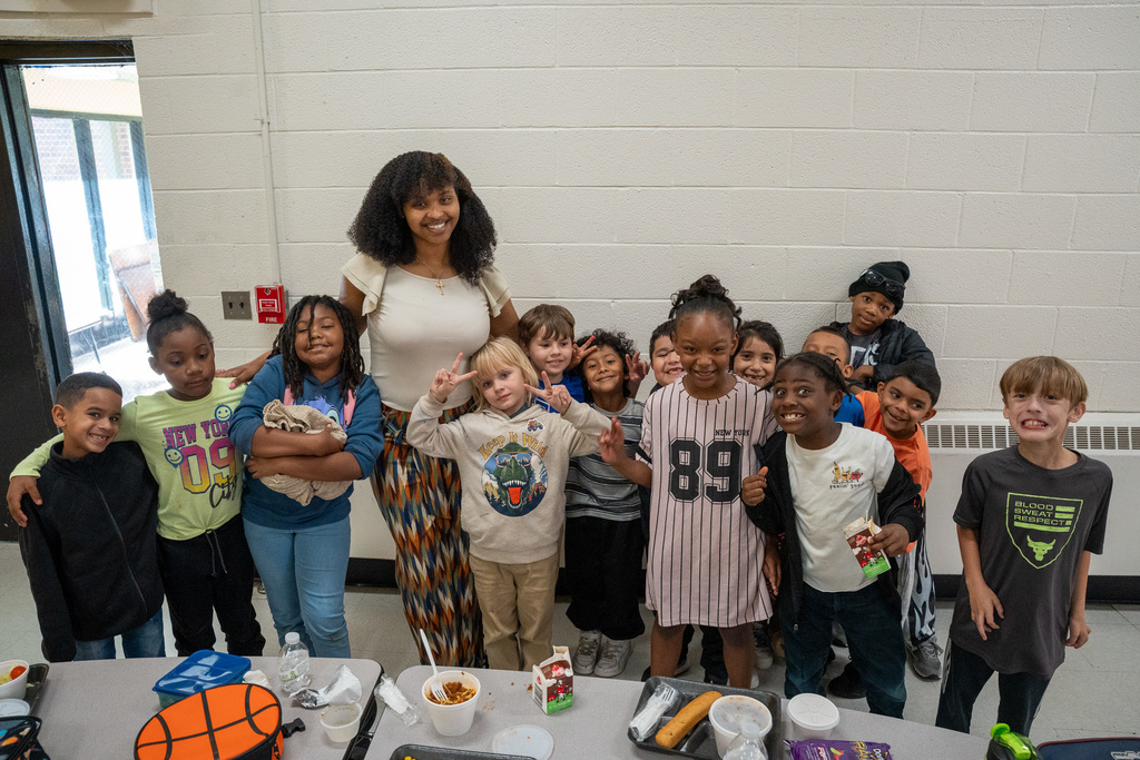 Large group of diverse elementary students gathered with teacher behind lunch table, all smiling at camera