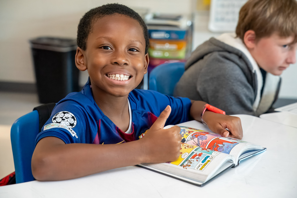 A young Black student in a blue soccer jersey smiles brightly at the camera while holding a marker over a comic book or graphic novel, sitting at a desk in a classroom with other students working in the background.