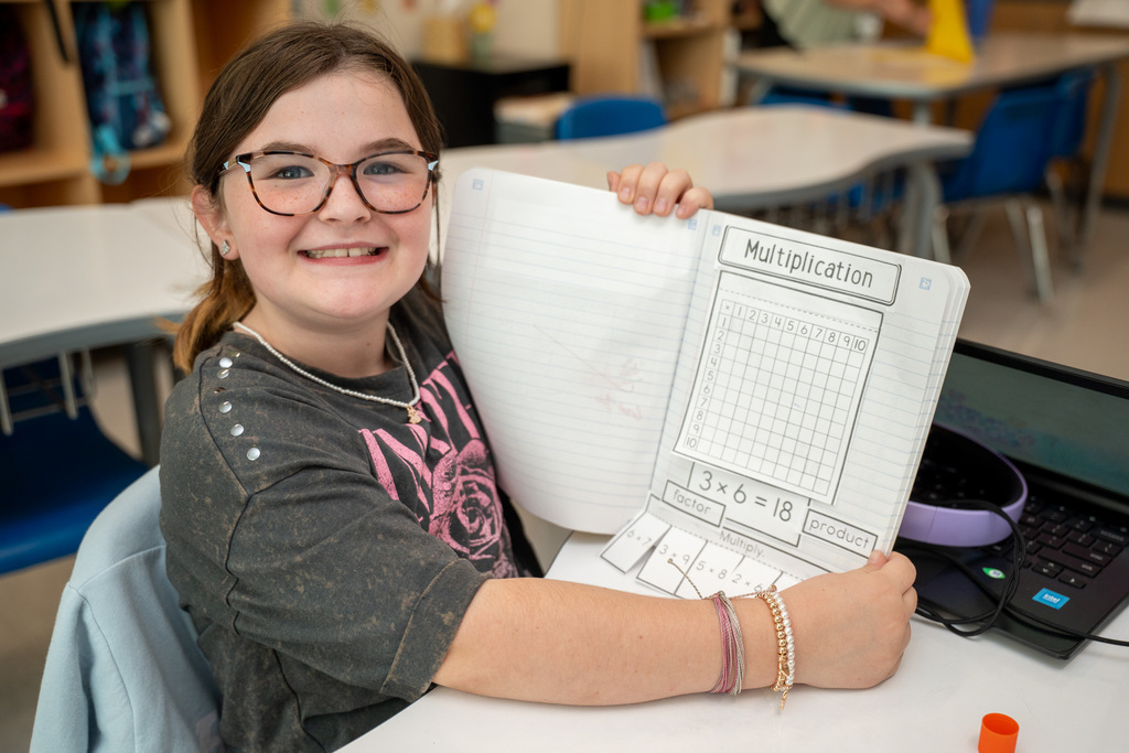 A young girl with glasses and bracelets holds up a multiplication worksheet showing a grid for practicing times tables, smiling at the camera while seated at a classroom desk with a laptop nearby.