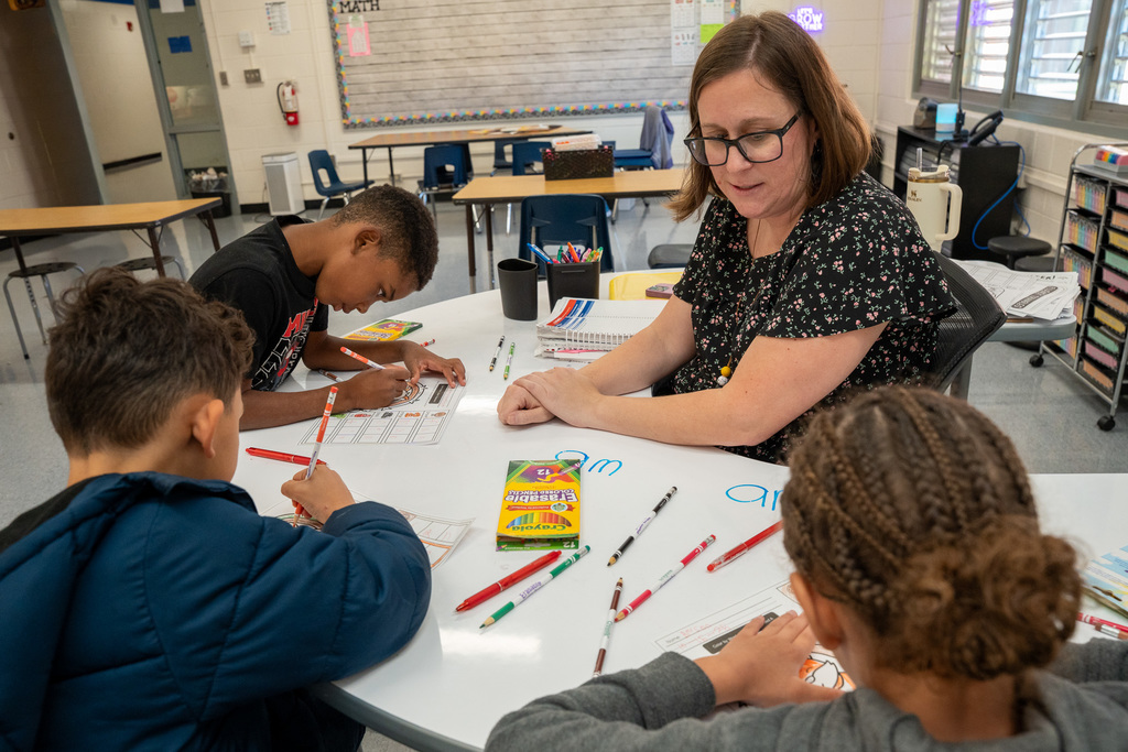 A teacher wearing glasses and a floral print dress sits at a white table with three elementary students in a classroom, working on math worksheets with colorful markers spread across the table. Math displays and student work are visible on the walls in the background.