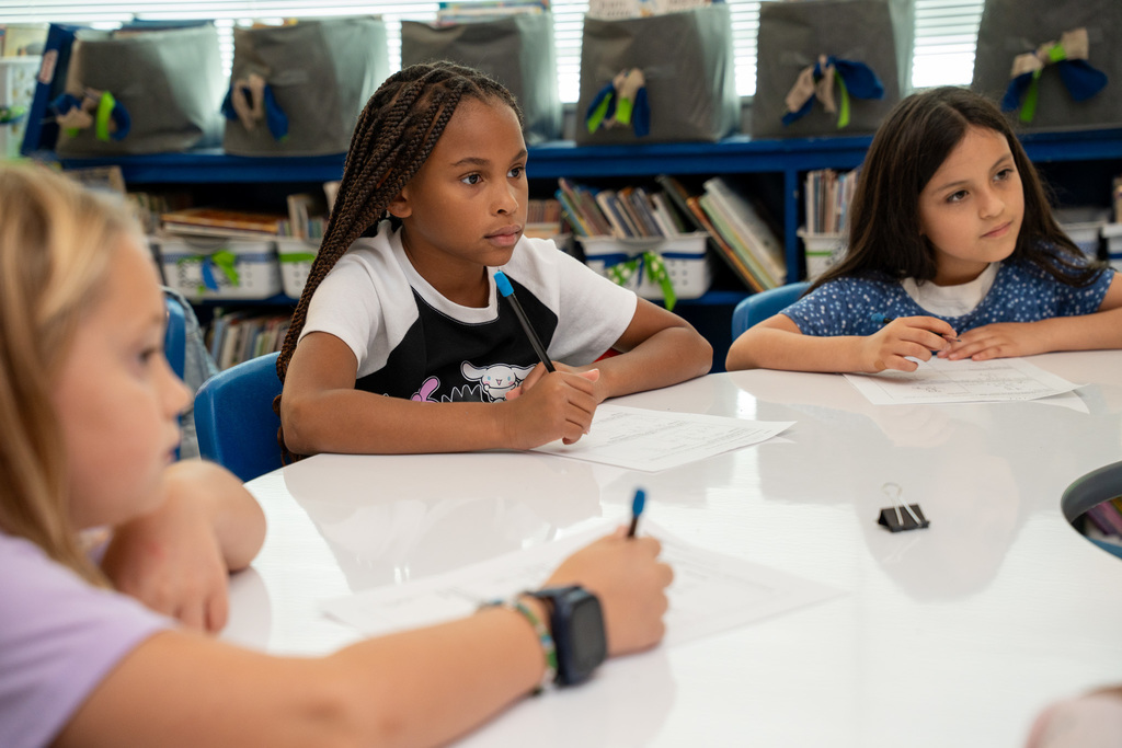 Four elementary students sit at a white table working on assignments, with a student in a black and white graphic tee centered in the frame, focused intently on their work. Classroom storage bins and materials are visible on shelves behind them.