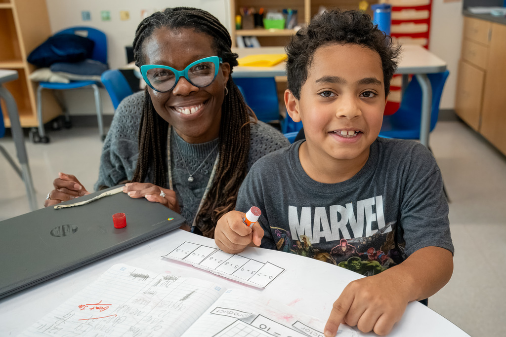 A teacher wearing distinctive turquoise cat-eye glasses sits next to a young boy in a Marvel t-shirt at a desk, both working on math worksheets with manipulatives and materials visible on the table.