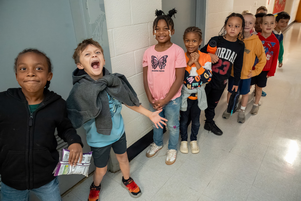 A group of enthusiastic elementary students line up in a school hallway, with two students in front making excited, silly faces at the camera while others smile and wait patiently behind them.