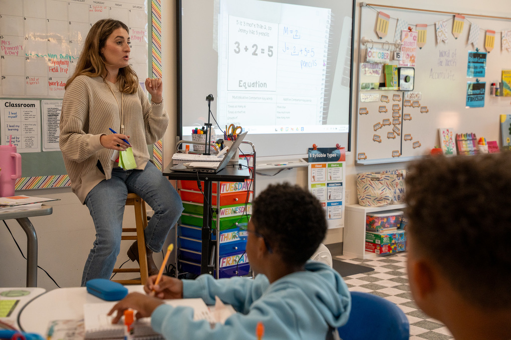 A teacher in a beige sweater sits on a desk at the front of a classroom, gesturing while teaching a math lesson about equations that's displayed on a projector screen behind her, with students seated at tables facing her.