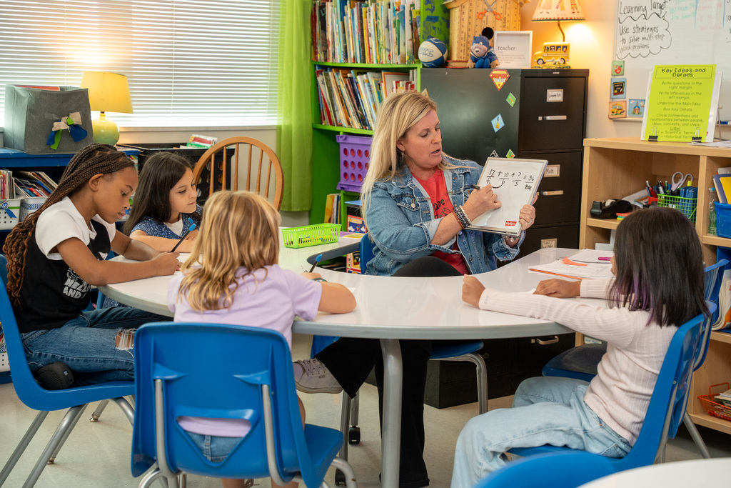 A teacher in a denim jacket sits at a small group table with five elementary students, holding up a whiteboard with a math problem while teaching. Books, pencil holders, and classroom materials surround the cozy learning space.