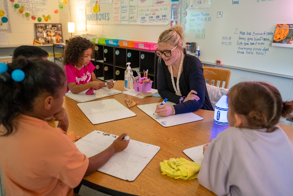 A teacher with her hair in a bun and wearing glasses works with a small group of elementary students at a wooden table, all focused on writing and math work. A whiteboard with math problems and colorful decorations are visible in the background.