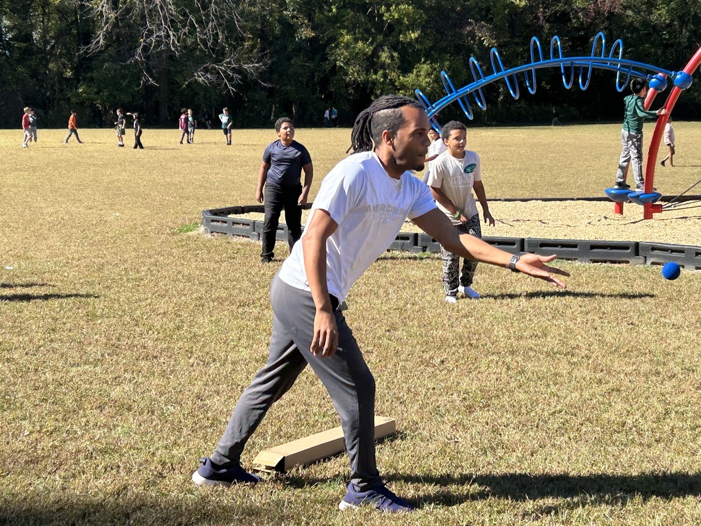 A young person throws a ball while playing on a sunny field during recess or outdoor activities, with other students and playground equipment visible in the background and a colorful climbing structure on the right side of the frame.