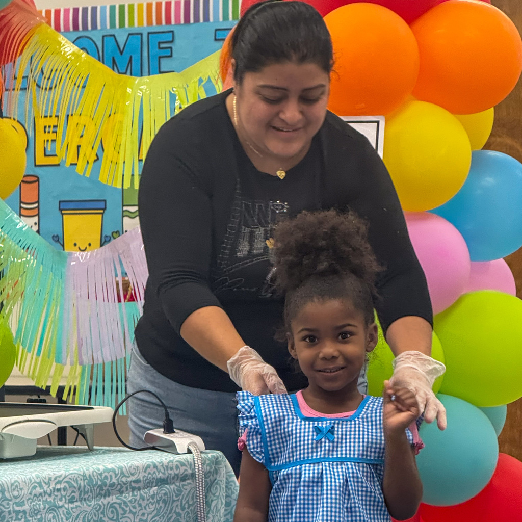 An adult woman in a black t-shirt with food service gloves helps a young girl wearing a blue gingham dress. The girl smiles at the camera while the woman guides her hands from behind. Rainbow balloons and streamers create a festive setting behind them.