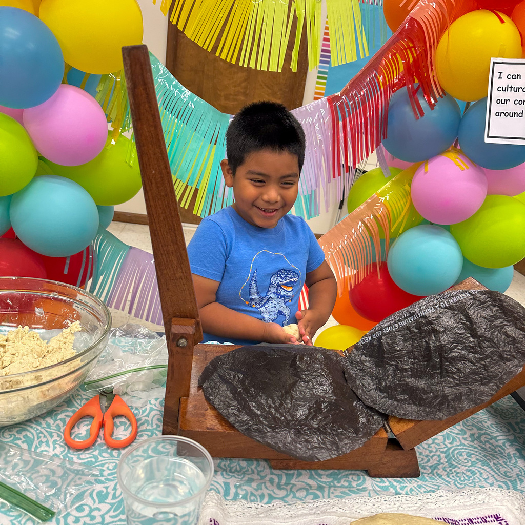 A young boy with short dark hair wearing a blue dinosaur t-shirt sits at a table working with the tortilla press. He smiles while handling dough. Orange scissors sit on the table corner near a bowl of masa and a glass of water. Rainbow balloon garland and colorful streamers decorate the background.
