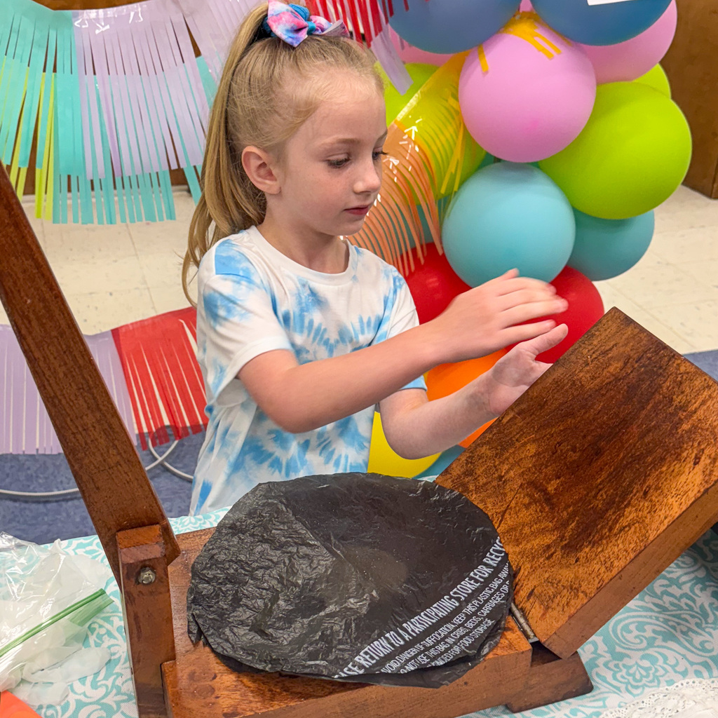 A young girl with blonde hair in a high ponytail wearing a tie-dye t-shirt uses both hands to operate a wooden tortilla press. She looks down concentrating on her work. The press sits on a table with finished tortillas, and rainbow balloons form an arch behind her.