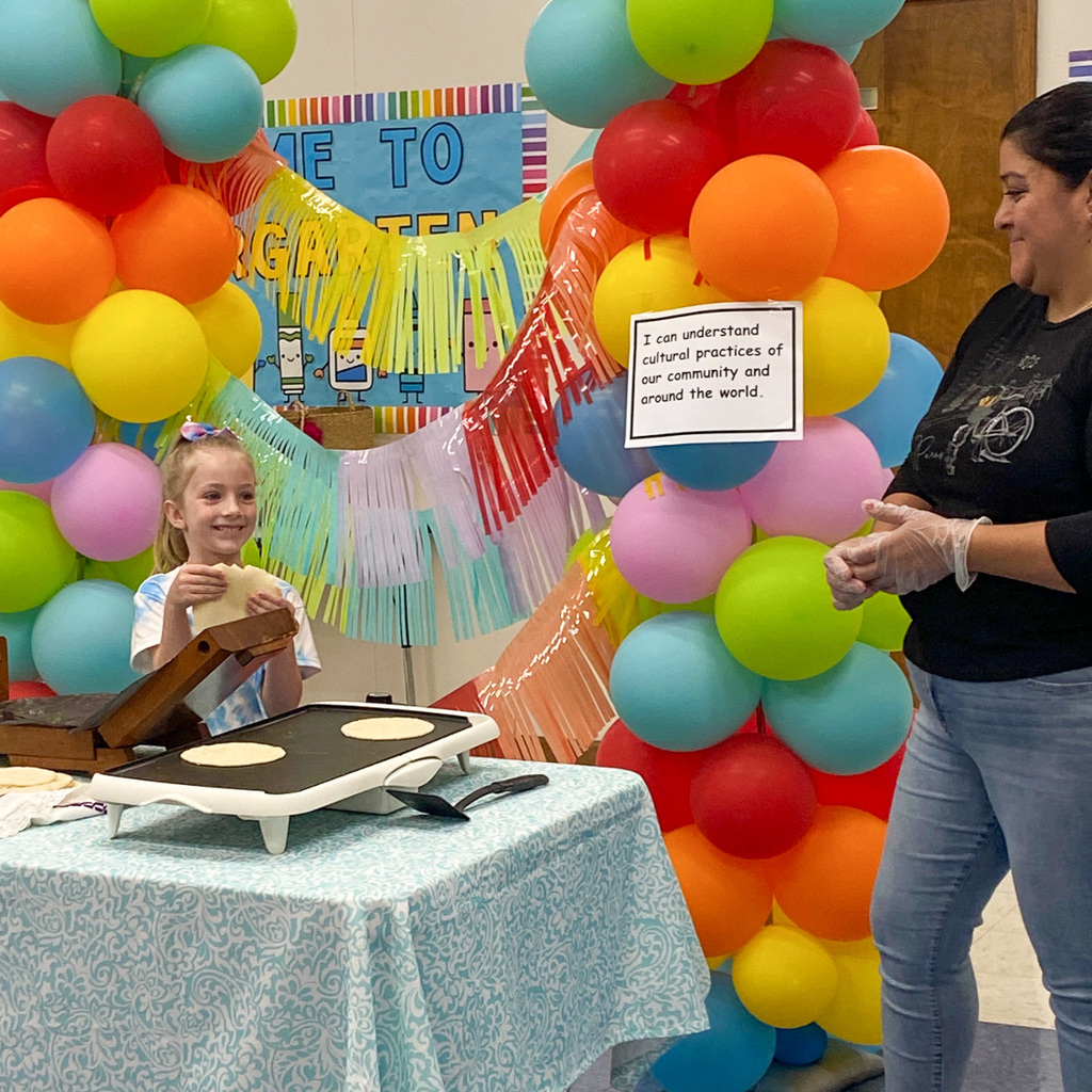 A young girl with blonde hair in a ponytail stands at a table with a tortilla press, smiling at the camera. An adult woman in a black t-shirt stands nearby wearing food service gloves. The background features a vibrant rainbow balloon arch with colorful fringe streamers and a sign reading "I can understand cultural practices of our community and around the world."