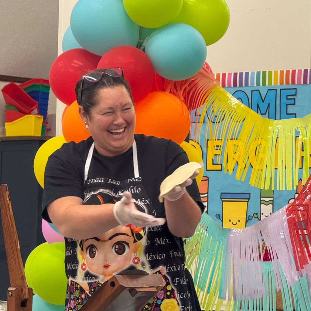 A woman with dark hair pulled back, wearing a black t-shirt with text about tortillas and Mexico, laughs heartily while holding a fresh tortilla in both gloved hands. Rainbow balloons create a colorful backdrop, and a sign partially visible behind her reads "WELCOME TO KINDERGARTEN."