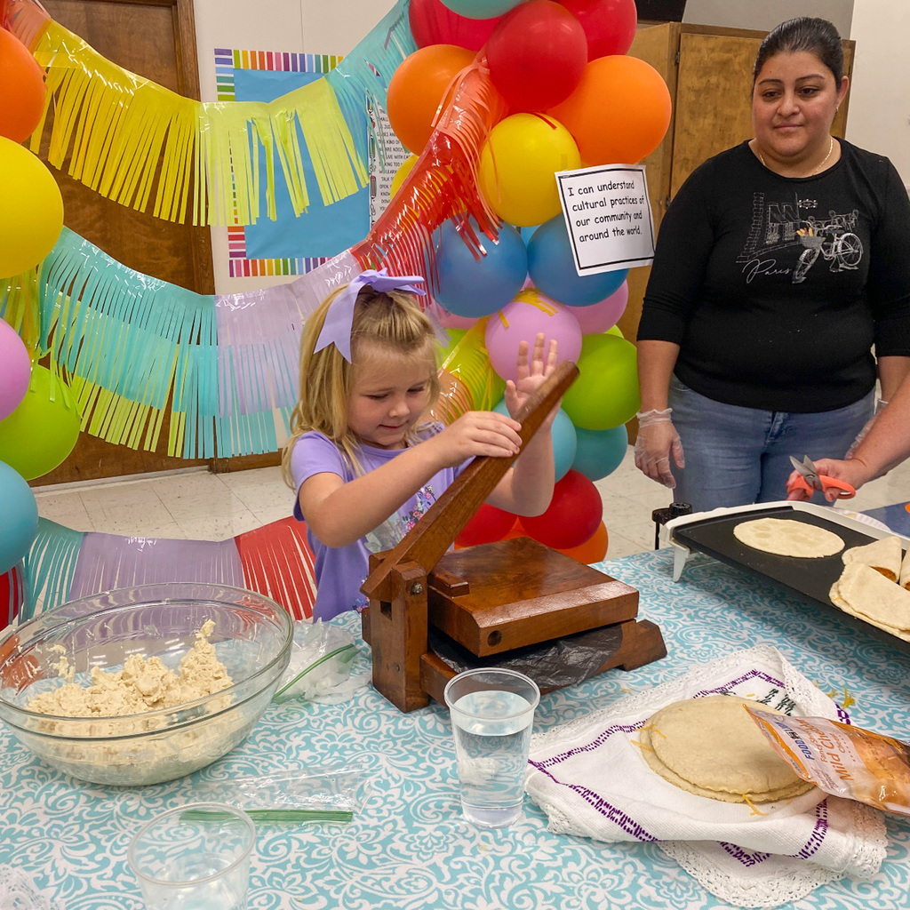 A young girl with blonde hair wearing a purple shirt uses a wooden tortilla press on a table. An adult woman in black stands beside her holding scissors near a griddle with tortillas. The table displays a bowl of masa dough, glasses of water, and finished tortillas on an embroidered cloth. Rainbow balloons and streamers decorate the background.