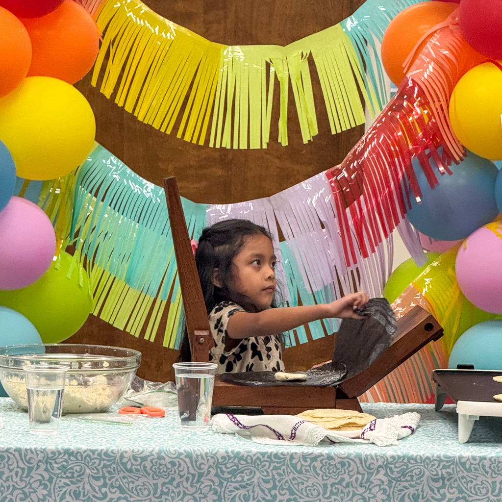 A young girl with dark hair in a ponytail wearing an animal print shirt operates a wooden tortilla press at a demonstration table. She looks upward while pressing. A bowl of masa dough, water glass, and finished tortillas are visible on the table. Colorful balloon garland and fringe decorations fill the background.