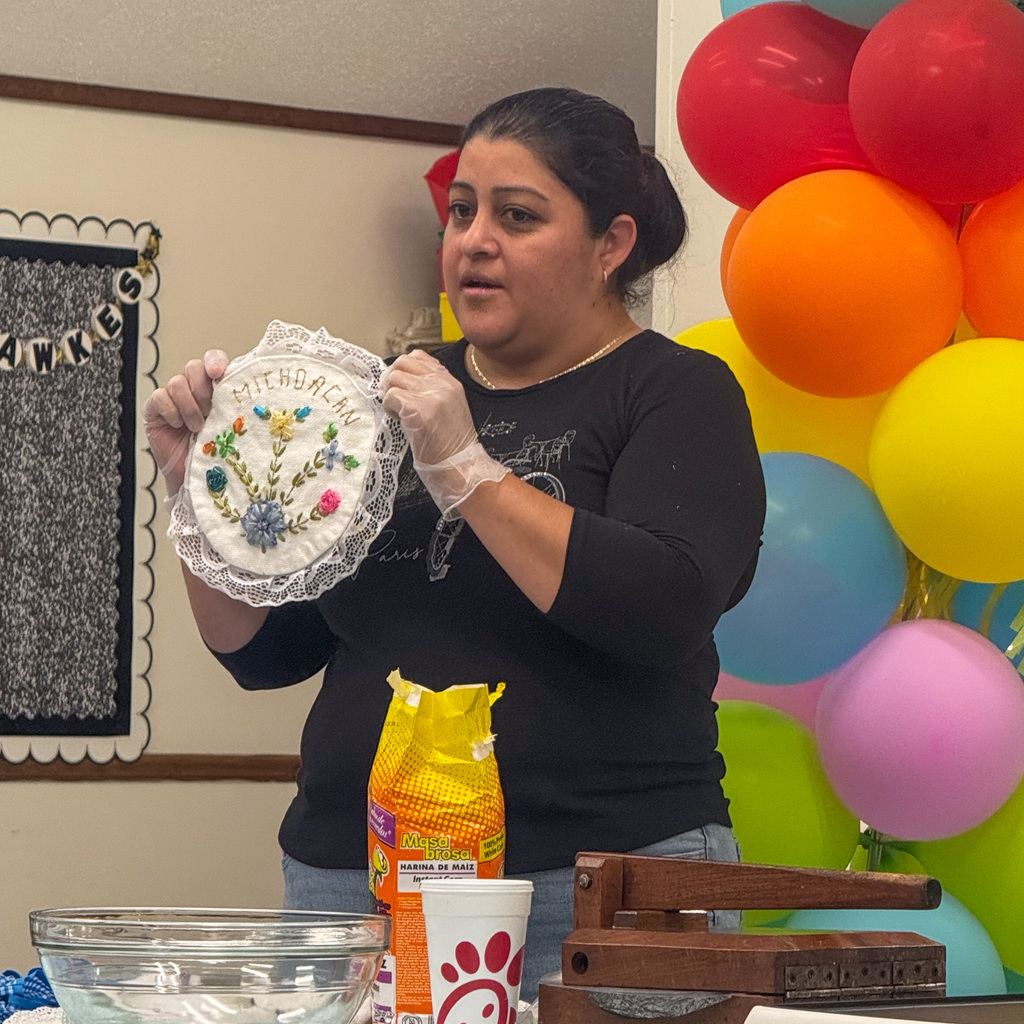 A woman with dark hair wearing a black long-sleeve shirt holds up an embroidered white cloth with lace trim displaying the word "MICHOACÁN" surrounded by colorful floral embroidery. She stands at a table with a bowl, a bag of Maseca corn flour, and a container visible. Rainbow balloons decorate the background.