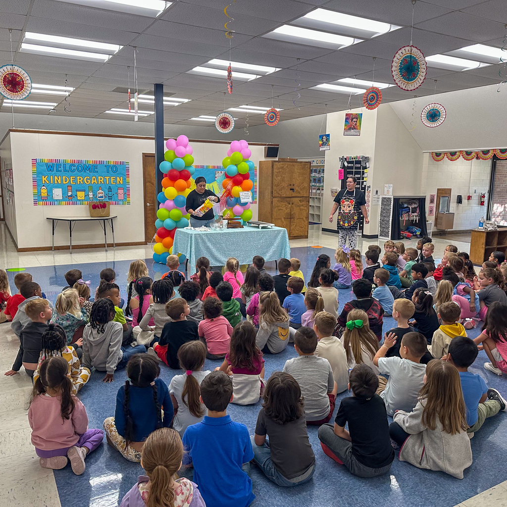 Wide view of a kindergarten classroom where children sit cross-legged on the floor in a large group, facing a demonstration table with a rainbow balloon arch. Two adults stand at the table conducting a presentation. A "Welcome to Kindergarten" sign is visible on the wall, along with decorative paper flowers hanging from the ceiling.