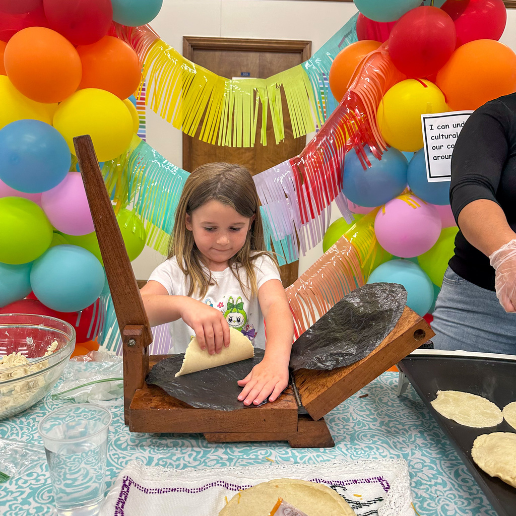 A young girl with long brown hair wearing a white t-shirt with a character design uses a wooden tortilla press, placing dough onto the press surface. The table shows a bowl of masa, water glasses, and finished tortillas on an embroidered cloth. Rainbow balloon garland and colorful fringe streamers create a festive backdrop.