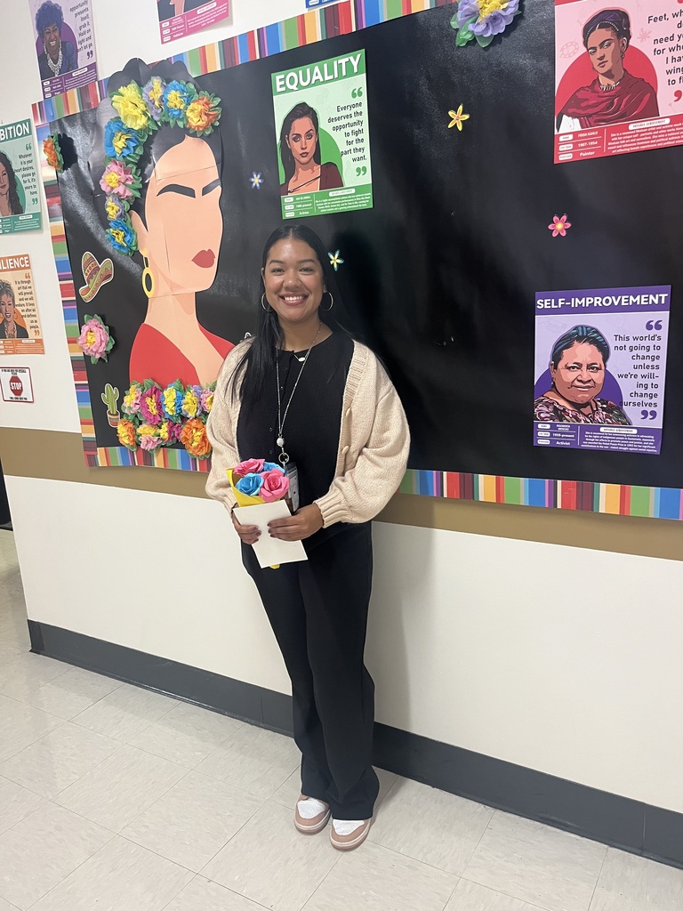 A young teacher wearing a black shirt and pants with khaki sweater stands in front of a bulletin board themed around Hispanic Heritage.
