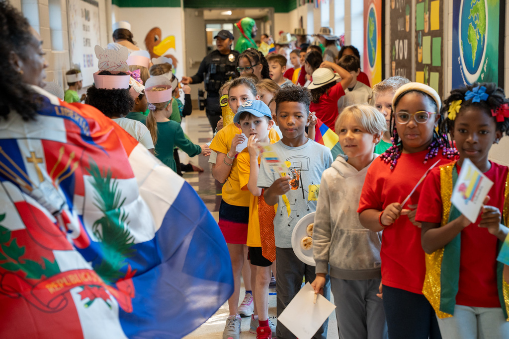 A diverse group of children parade through a school hallway carrying various flags, including the Dominican Republic flag prominently displayed in the foreground. Students wear different colored clothing and accessories representing various countries while spectators line the hallway.  Image 7: A large group of young students poses with their teacher in front of a bulletin board reading "In a world where you can BE ANYTHING BE PREPARED." All the children hold small Dominican Republic flags and wear coordinating red, white, and blue colors or ribbons.  Image 8: A teacher in traditional Dominican dress with a parrot headpiece poses with students wearing animal masks representing Costa Rican wildlife. The children hold the Costa Rican flag featuring blue, white, and red horizontal stripes, and many wear green clothing.  Image 9: A classroom of diverse students displays the Colombian flag while wearing various cultural accessories and holding papers. Behind them is a colorful bottle cap mural depicting Earth with continents, and a teacher stands with them in a Colombia t-shirt.  Image 10: Students of various ages pose with the Colombian flag in front of a "BE ANYTHING BE PREPARED" bulletin board. They wear traditional Colombian hats (sombreros vueltiaos), hold small Colombian flags, and some wear costumes representing different aspects of Colombian culture.