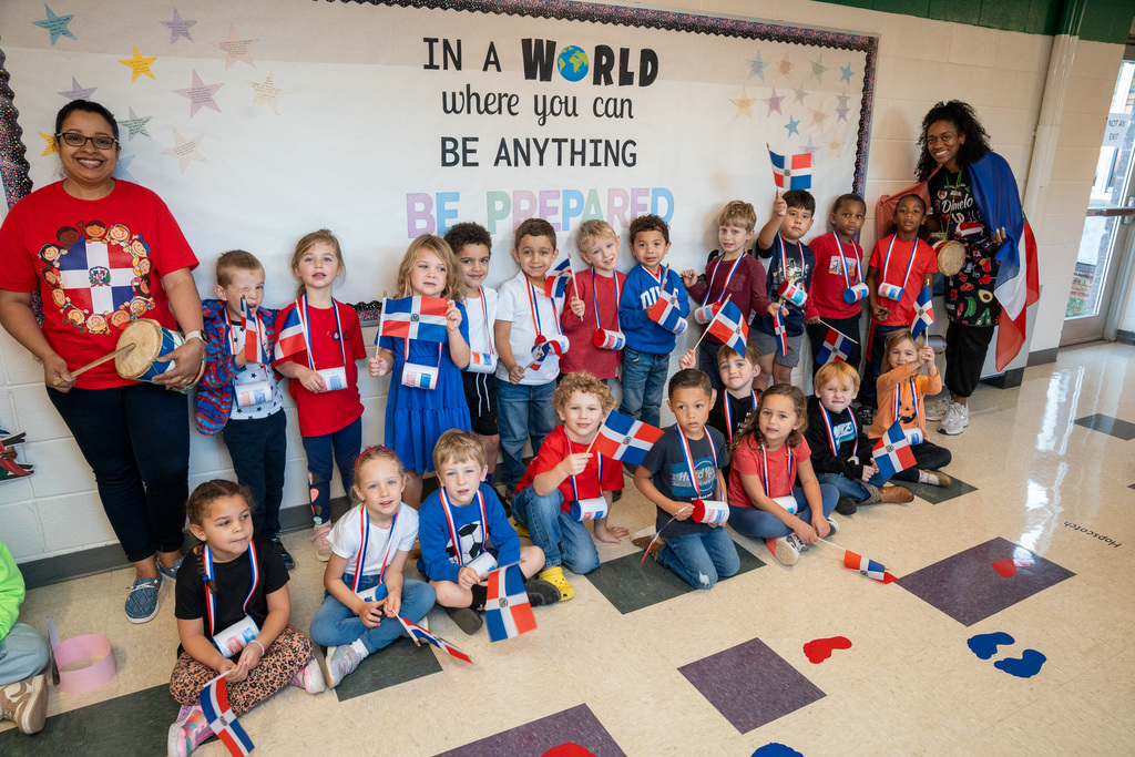 A large group of young students poses with their teacher in front of a bulletin board reading "In a world where you can BE ANYTHING BE PREPARED." All the children hold small Dominican Republic flags and wear coordinating red, white, and blue colors or ribbons.
