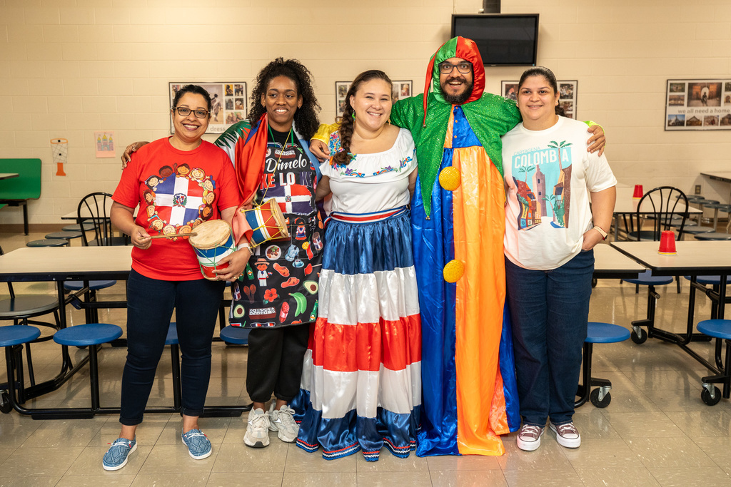 Five adults pose together in a school cafeteria wearing clothing representing different Latin American countries. They display traditional attire including Dominican Republic t-shirts, a traditional Dominican dress, and a colorful Colombian-style costume, with one person holding a drum.