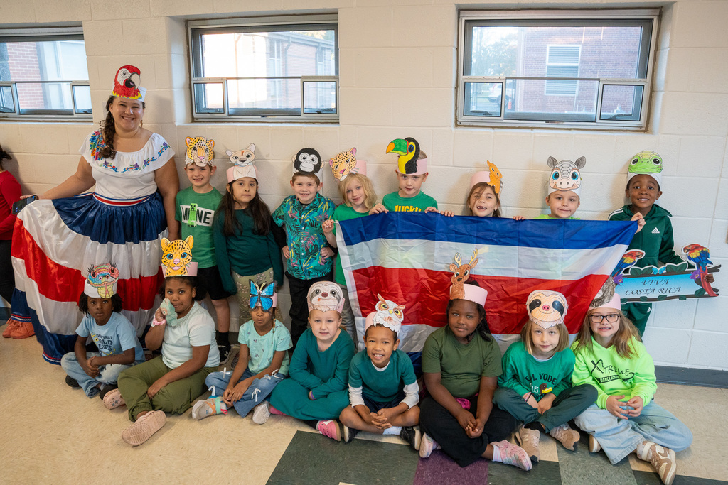 A teacher in traditional Dominican dress with a parrot headpiece poses with students wearing animal masks representing Costa Rican wildlife. The children hold the Costa Rican flag featuring blue, white, and red horizontal stripes, and many wear green clothing.