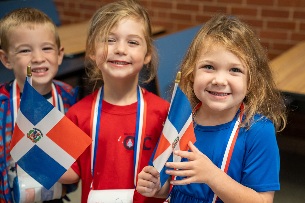 Three young children wearing Dominican Republic flag colors smile at the camera while holding small flags. They're dressed in red and blue with red-white-and-blue striped ribbons around their necks, standing in front of a brick wall in what appears to be a school cafeteria.