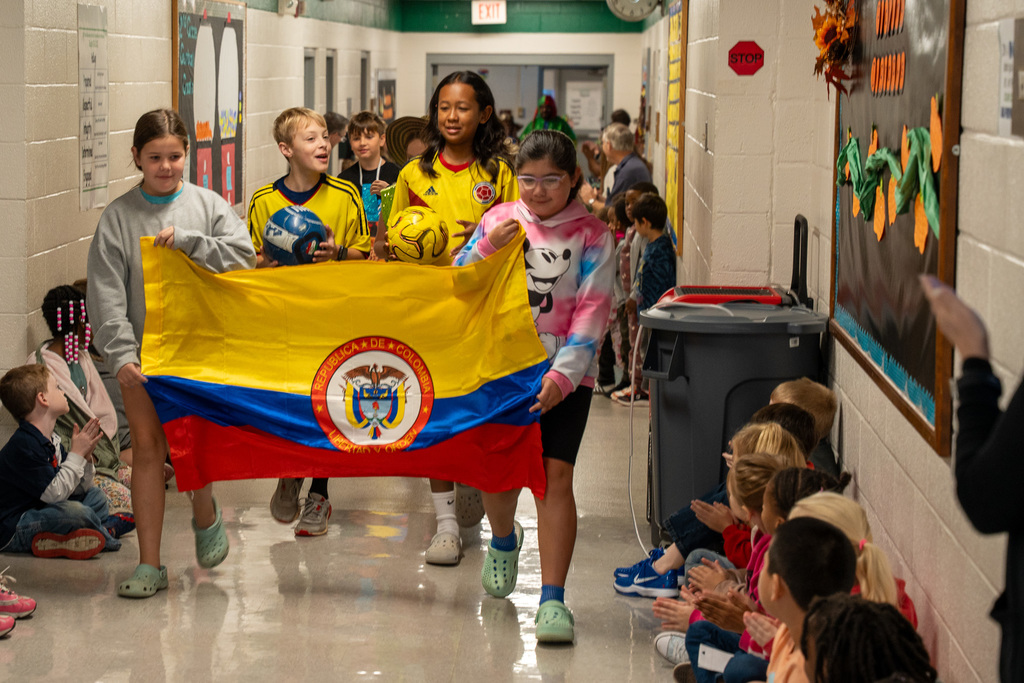 Students parade through a school hallway carrying the Colombian flag with its distinctive yellow, blue, and red horizontal stripes and national coat of arms. Children wearing Colombian soccer jerseys lead the procession while other students sit along the hallway walls watching.