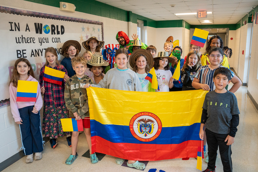Students of various ages pose with the Colombian flag in front of a "BE ANYTHING BE PREPARED" bulletin board. They wear traditional Colombian hats (sombreros vueltiaos), hold small Colombian flags, and some wear costumes representing different aspects of Colombian culture.