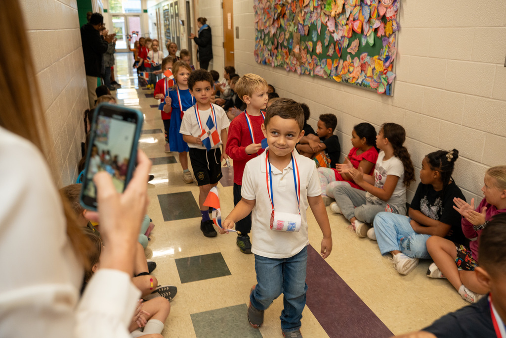 Young children walk in a school hallway parade wearing Dominican Republic colors and holding small Dominican flags on sticks. The students have red, white, and blue ribbons around their necks while other children sit along the walls watching the celebration.