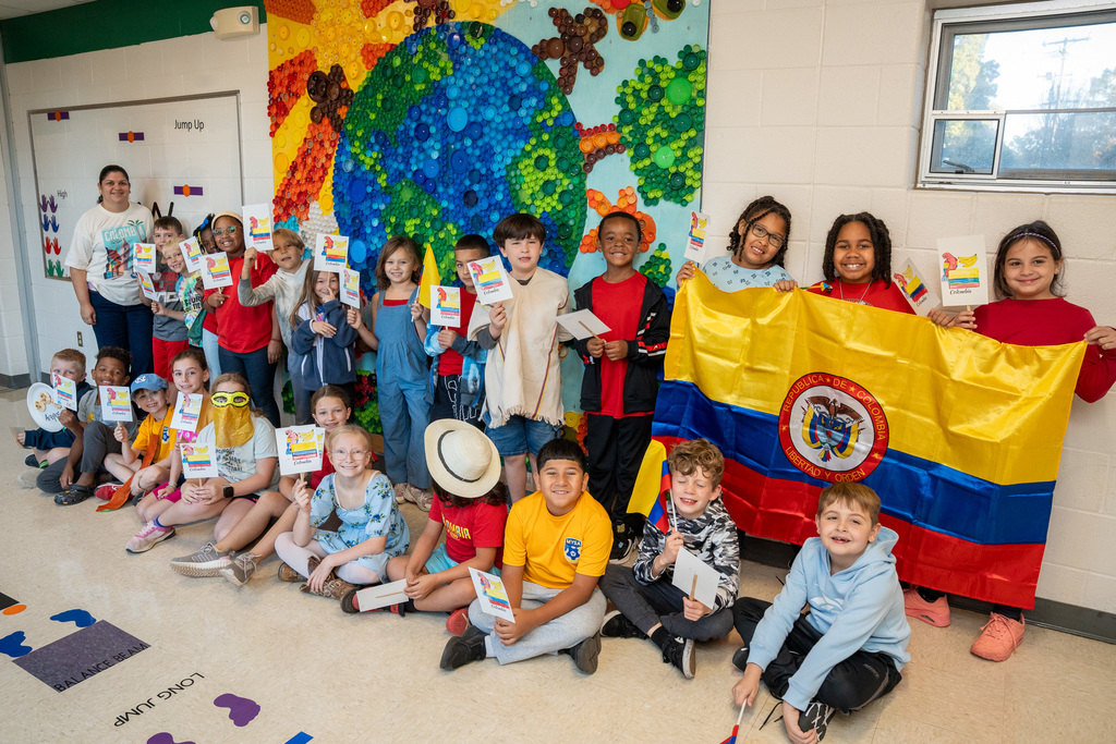 A classroom of diverse students displays the Colombian flag while wearing various cultural accessories and holding papers. Behind them is a colorful bottle cap mural depicting Earth with continents, and a teacher stands with them in a Colombia t-shirt.