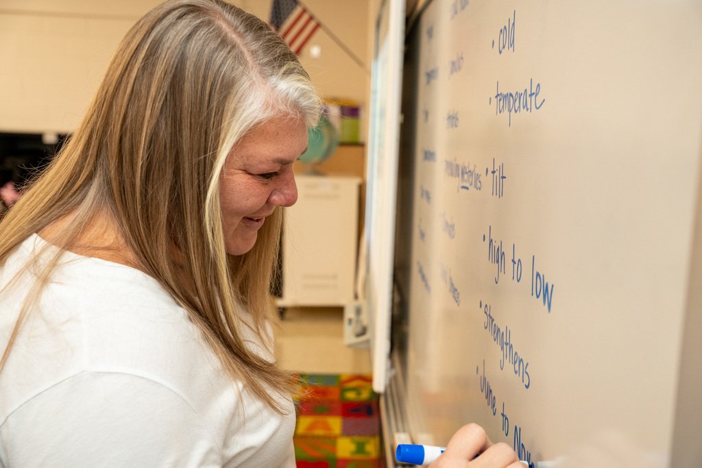 A woman with long blonde and gray hair wearing a white top writes on a whiteboard with a blue marker, smiling while writing educational content including words like "temperate" and "high to low."