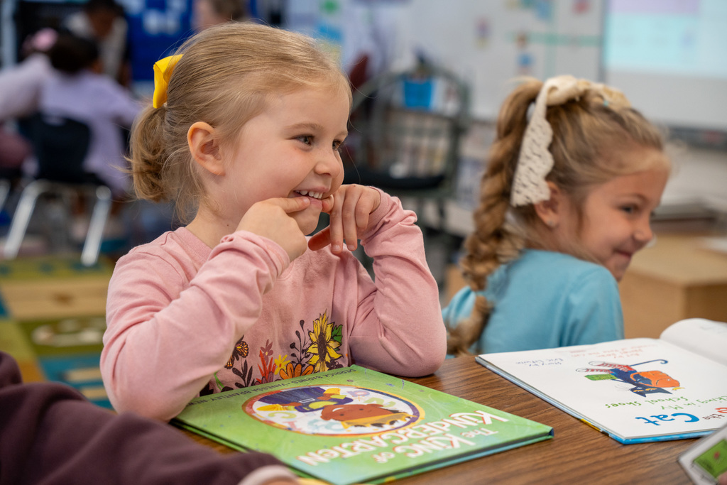 A young girl with blonde hair in a ponytail with a yellow bow, wearing a pink sweatshirt with floral design, sits at a classroom table smiling with her hand near her mouth. She has a green book in front of her while another student works beside her.