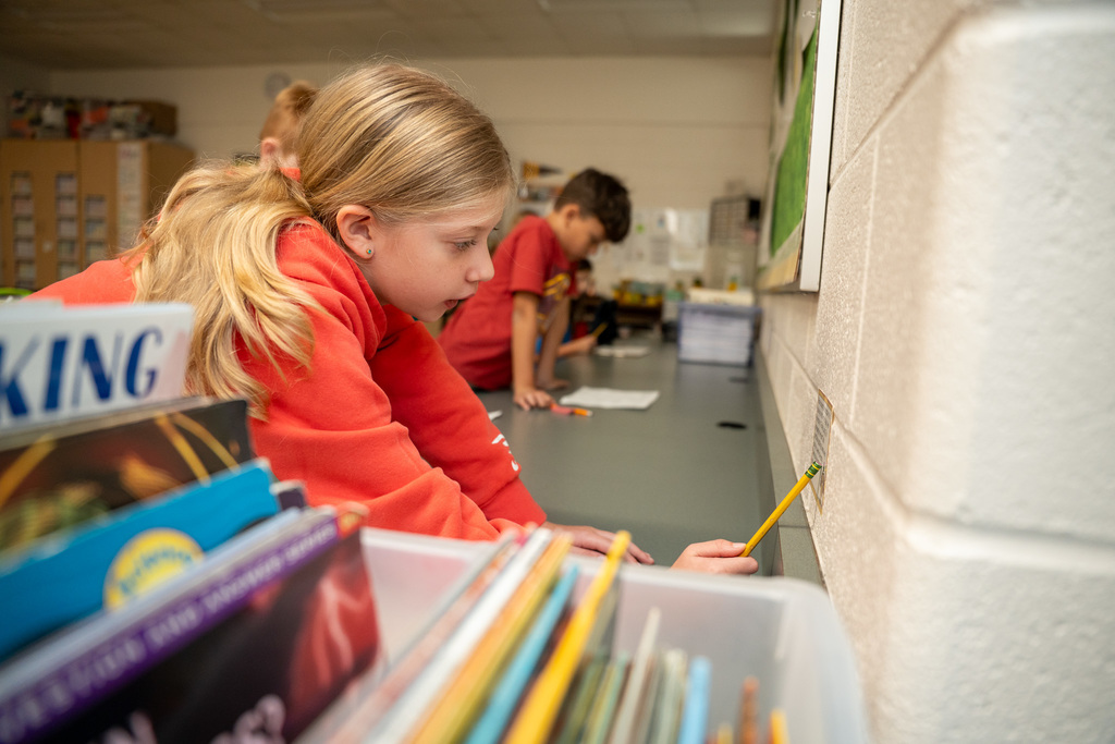 A young girl with blonde hair in pigtails wearing an orange hoodie leans forward at a desk, holding a yellow pencil and writing on paper while books are stacked nearby in a classroom setting.