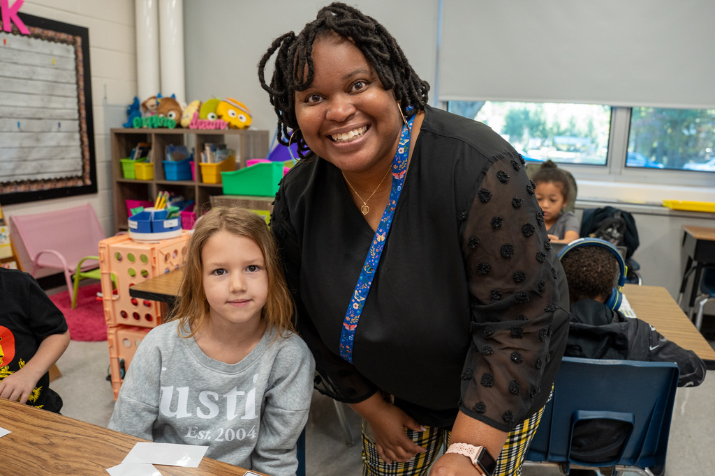A Black female educator with locs wearing a black top with textured details and a blue patterned lanyard poses with her arm around a young girl with long brown hair wearing a gray sweatshirt in a colorful classroom setting.