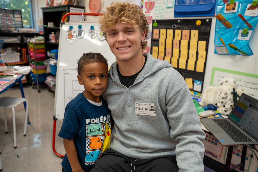 A young male visitor with curly blonde hair wearing a gray zip-up hoodie sits beside a young Black boy in a navy Pokémon t-shirt, both smiling at the camera in a classroom filled with learning materials and bulletin boards.