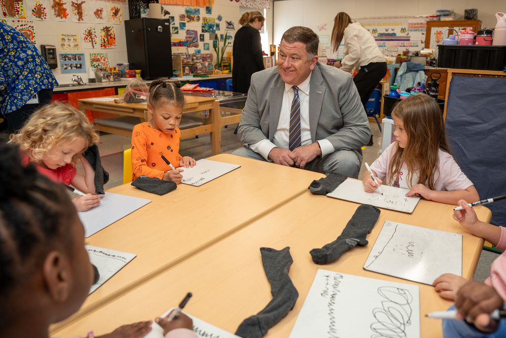 A man in a gray suit and striped tie sits at a round table with young students, smiling while children work on papers, with pairs of gray socks placed on the table surface in a classroom decorated with student artwork.