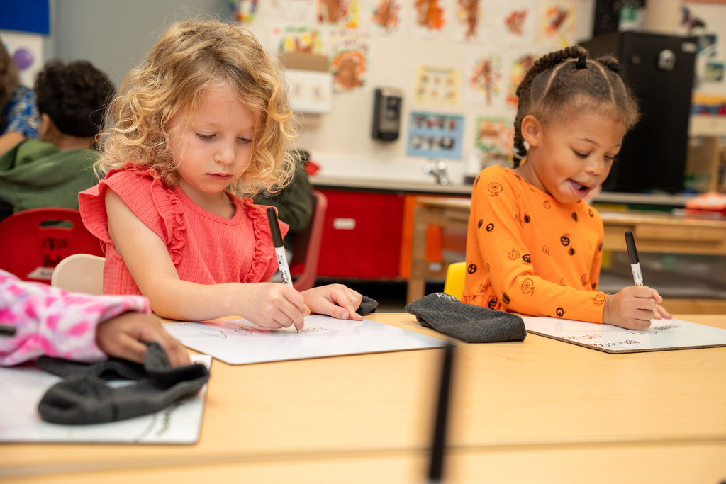Two young girls sit at a classroom table working on papers with markers - one with blonde curly hair wearing a coral pink ruffled top, and another with hair in a bun wearing an orange shirt with small prints, both focused on their work with gray socks on the table.