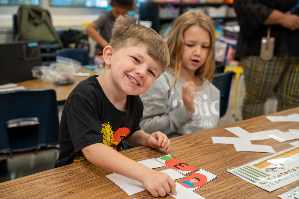A young boy with short brown hair wearing a black t-shirt with a graphic design sits at a wooden table smiling broadly at the camera, with educational materials and cards visible on the desk and a female student working beside him.