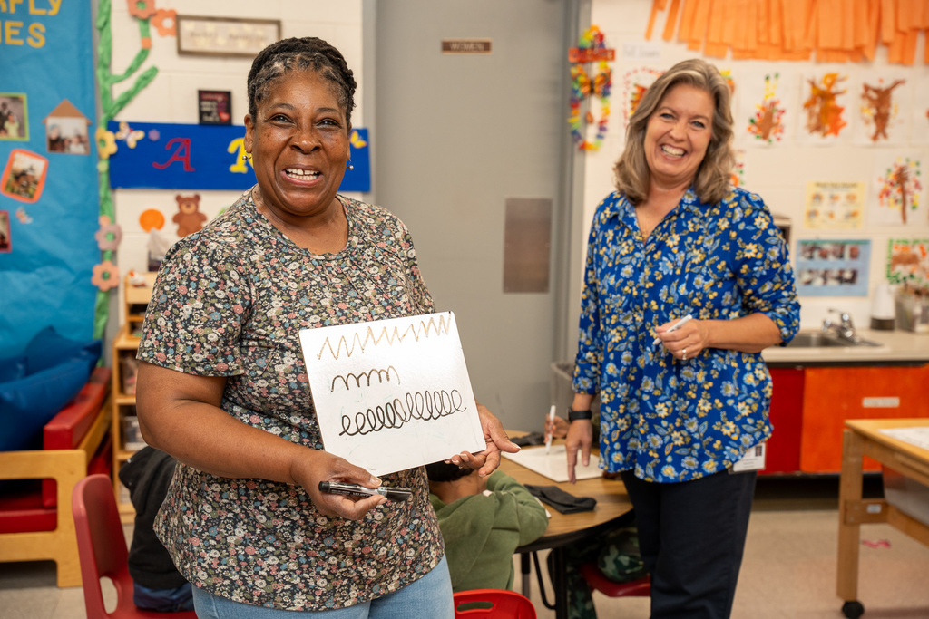 A Black female educator wearing a floral print top holds up a white paper displaying wavy line writing practice patterns, smiling broadly while another woman in a blue floral dress stands in the background in a classroom setting.