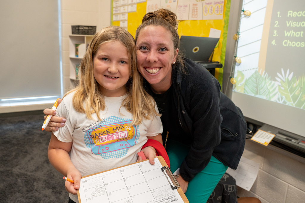 A woman with her hair in a bun wearing a black jacket and teal pants smiles while kneeling beside a young blonde girl in a graphic t-shirt who holds a clipboard and orange marker, both posing together in a classroom with a window and educational displays visible behind them.