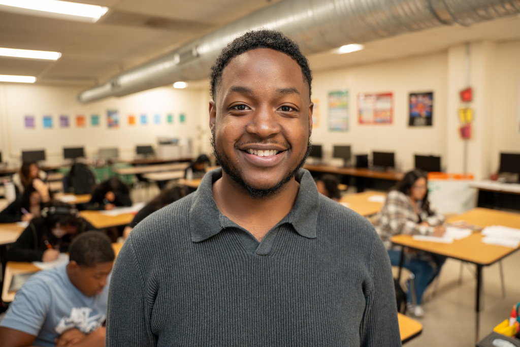 A close-up portrait of Brandon Daye, a teacher with a warm smile, standing in the middle of his classroom. Students are seated and working at desks in the background.
