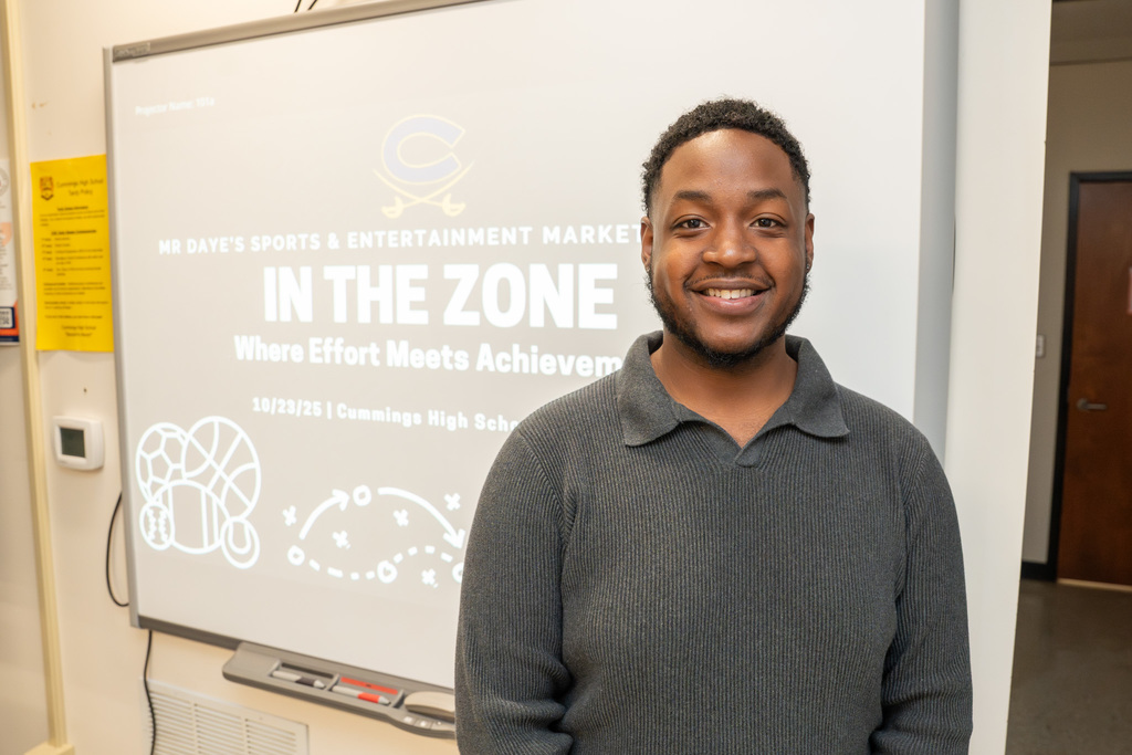 Sports Marketing teacher Brandon Daye stands in his classroom with seven students. Daye holds a basketball and smiles at the camera. The students hold items related to sports and marketing, including a 'Sports Marketing' binder and a mascot figurine.