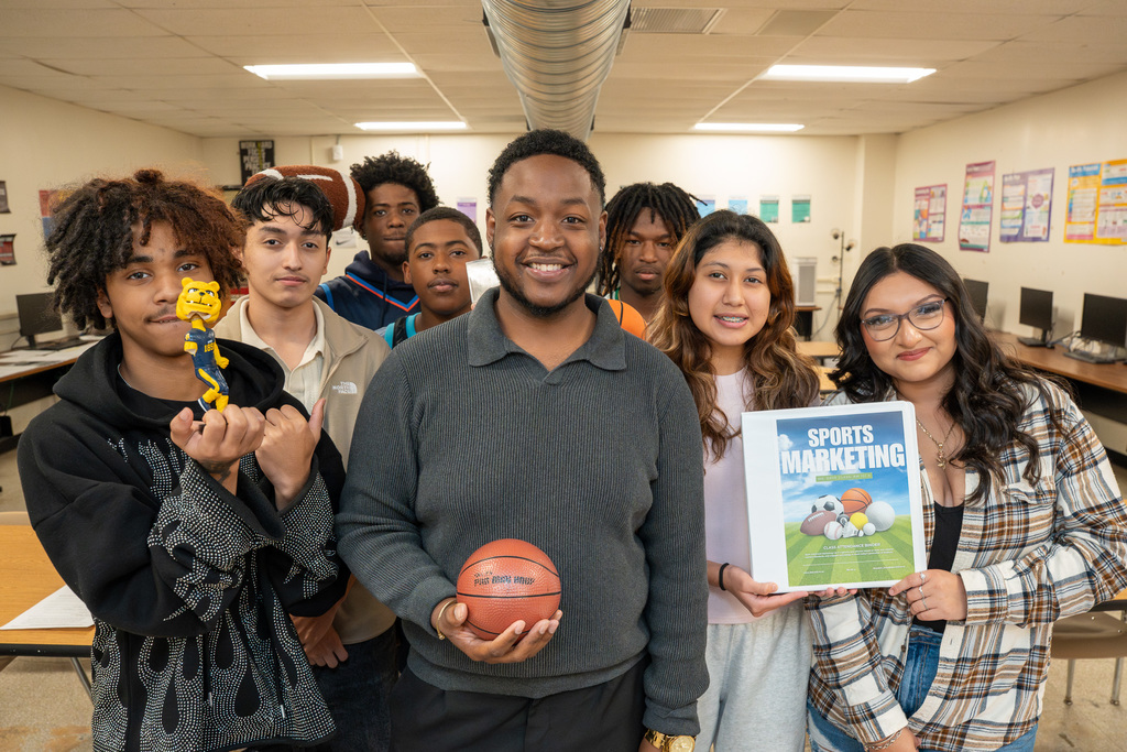 Brandon Daye stands smiling in his classroom in front of a white interactive whiteboard. The board displays a graphic for 'MR. DAYE'S SPORTS & ENTERTAINMENT MARKET[ING]' with the title 'IN THE ZONE: Where Effort Meets Achievement'.