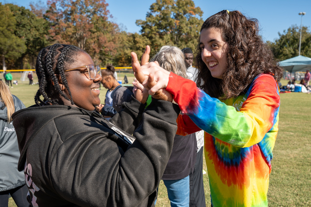 Two teenage girls, one in a black hoodie and the other in a vibrant tie-dye shirt, smile and joyfully slap hands (high-five) while standing outdoors.